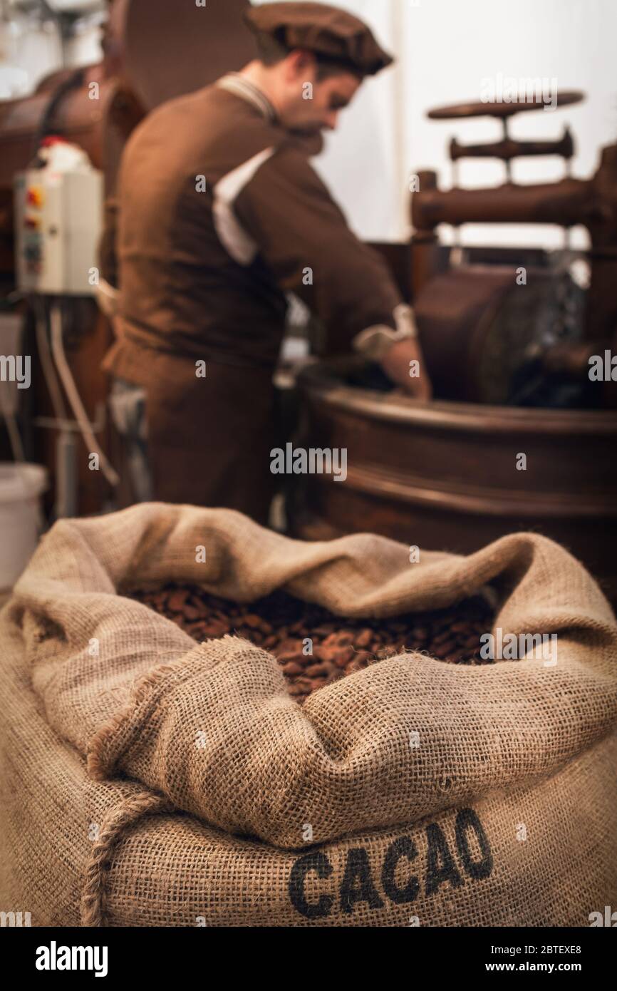 Borsa di iuta piena di chicchi di cacao in un laboratorio di cioccolateria, con un cioccolatiere maschile che lavora su attrezzature per conching e melanger sullo sfondo Foto Stock