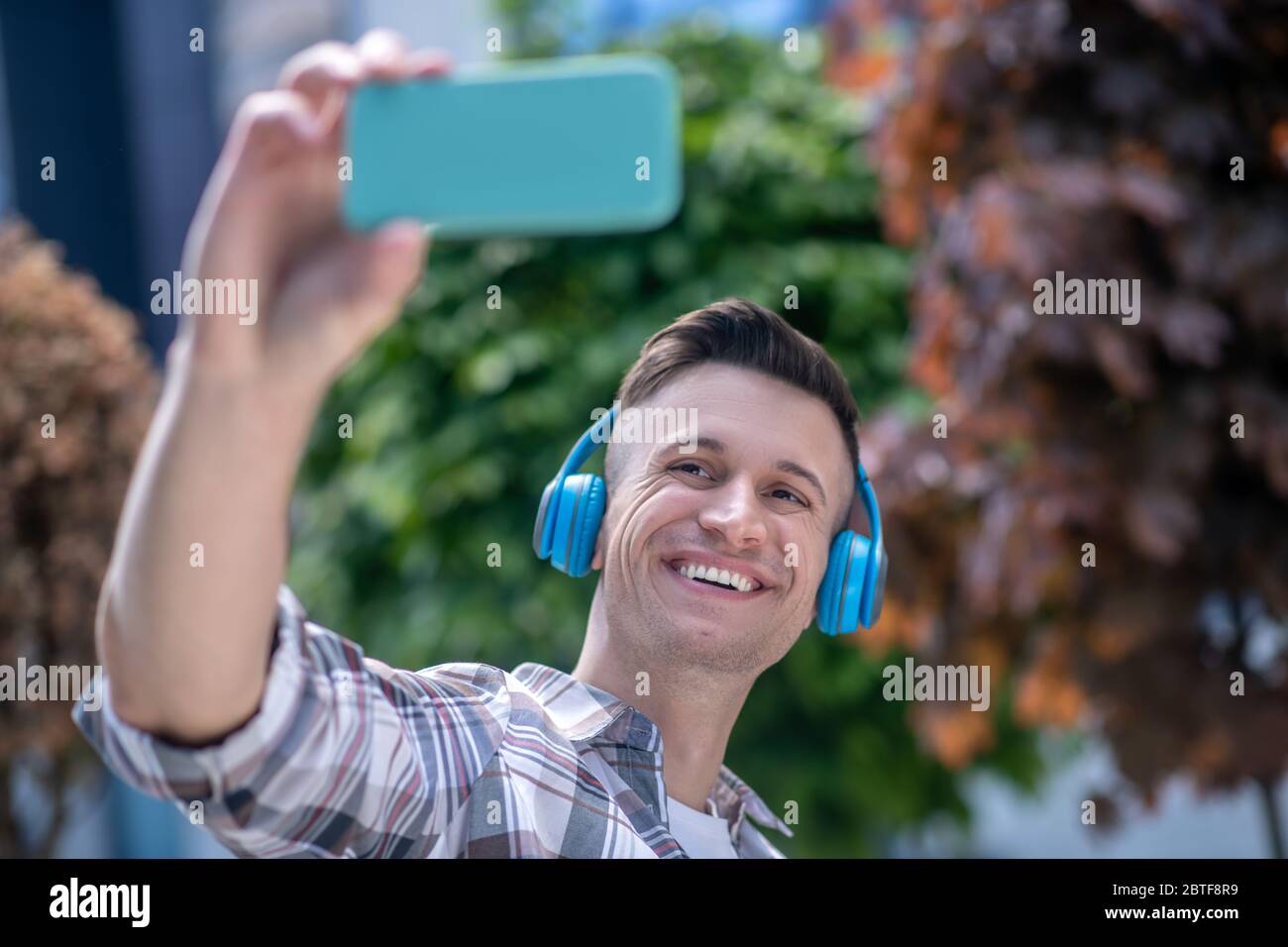 Allegro maschio dai capelli scuri che indossa le cuffie, facendo selfie, sorridendo Foto Stock