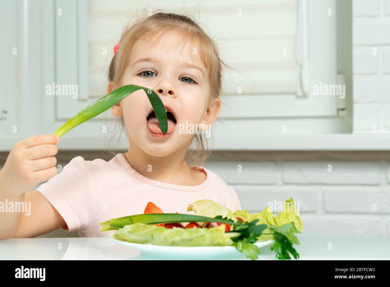 4 anni ragazza siede in cucina al tavolo e mangia verdure, morde cipolle verdi. preschool nutrizione regole Foto Stock