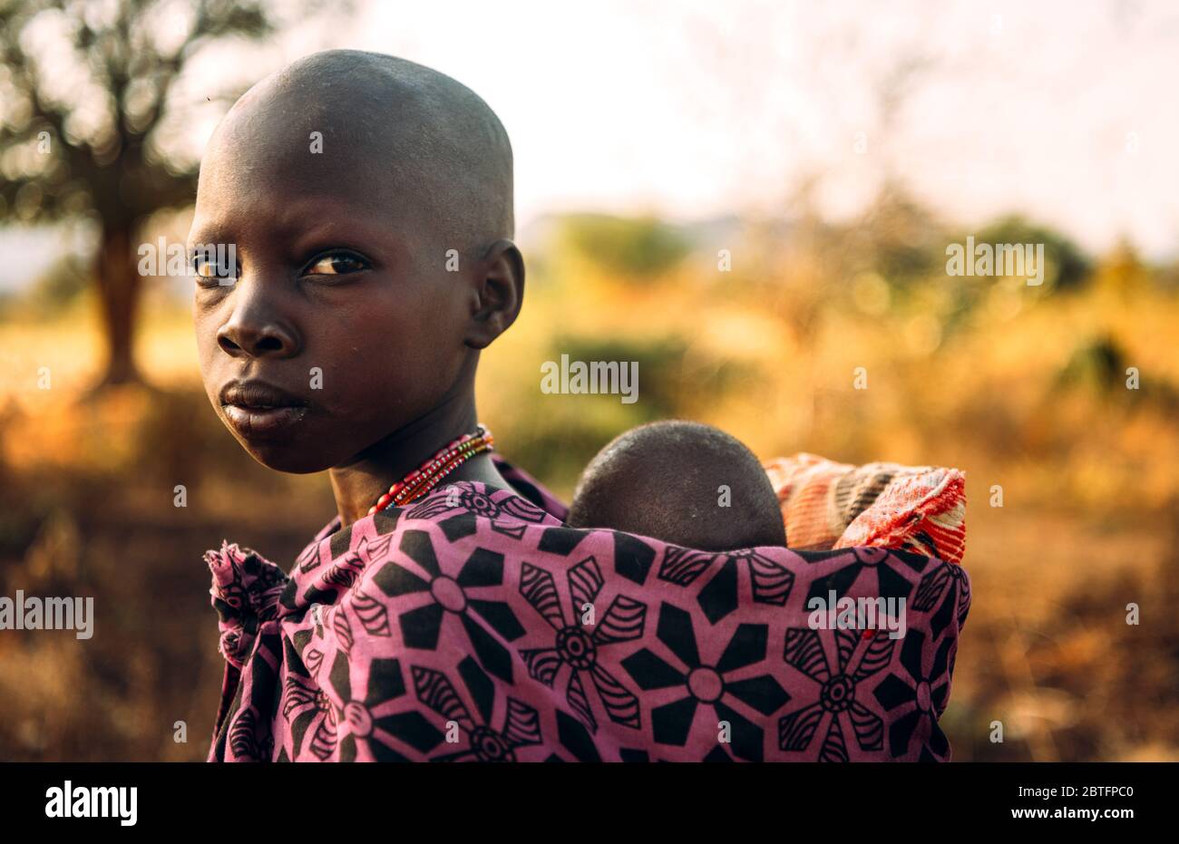 TRIBÙ BOYA, SUD SUDAN - 10 MARZO 2020: Ragazza in abito colorato tradizionale di Boya Tribe portare il bambino sulla schiena su sfondo sfocato di savana in Foto Stock