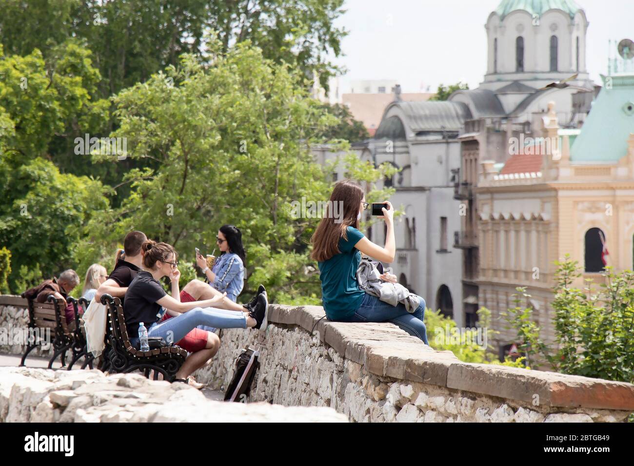 Belgrado, Serbia - 21 maggio 2020: Ragazza adolescente seduta su un muro della fortezza di Kalemegdan e filmare la vista della città con il telefono cellulare, e gruppo di giovani Foto Stock