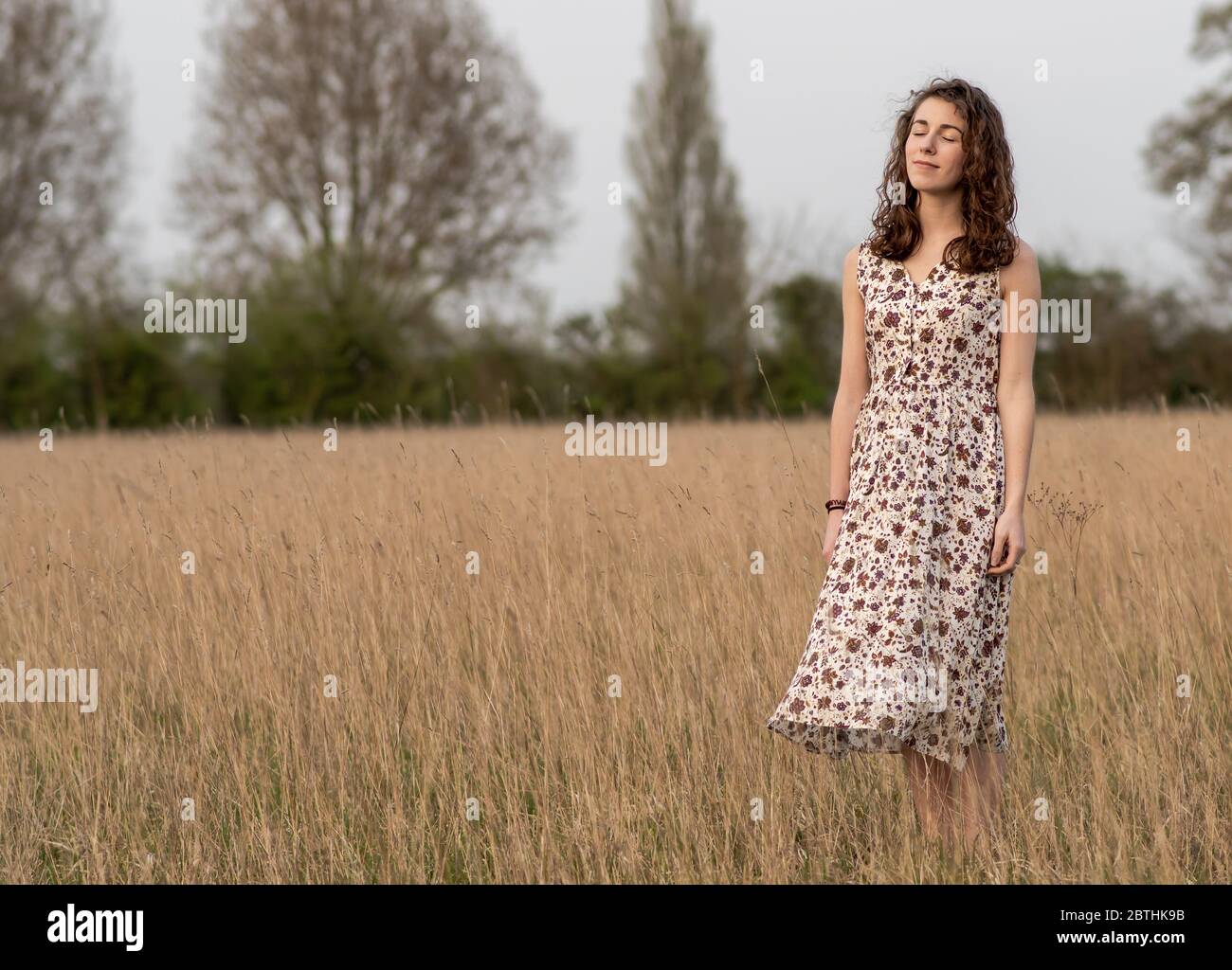 Una bella e sana giovane donna in un abito danze si trova pacificamente meditando e pensando in un campo di erba marrone al tramonto Foto Stock