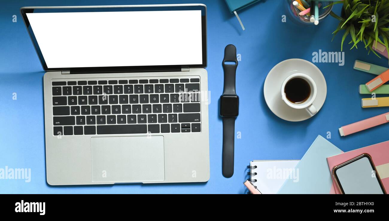 Immagine dall'alto del computer portatile con schermo bianco vuoto che si trova su una scrivania colorata circondata da orologio intelligente, caffè, notebook, smartphone Foto Stock