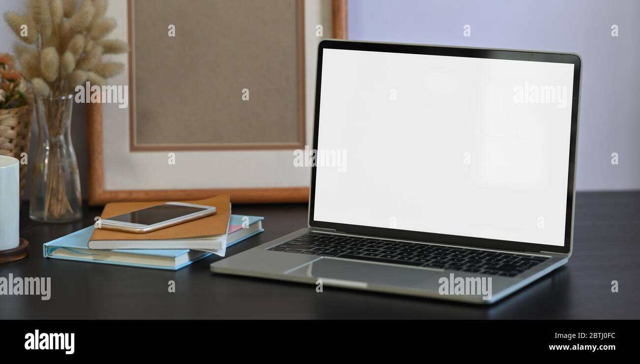 Foto di computer portatile bianco a schermo vuoto che si trova su una scrivania nera e circondato da una pila di notebook, smartphone, cornice vuota, jolly Foto Stock
