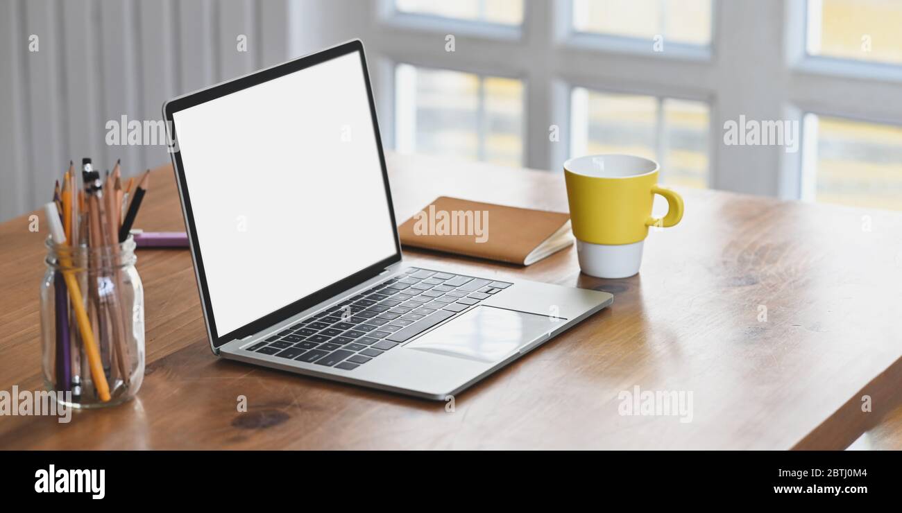 Computer portatile con schermo bianco vuoto che si mette su scrivania di legno e circondato da caffè tazza, nota, matite in vaso di vetro e pennarello sopra c Foto Stock