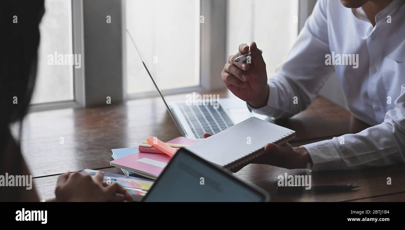 Immagine ritagliata di uomo d'affari prendendo appunti mentre si siede di fronte al computer portatile e il suo collega alla scrivania di legno sopra salotto vincere Foto Stock