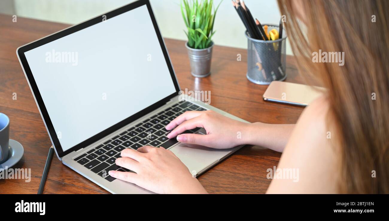Corto di bella donna che scrive su bianco schermo computer portatile bianco che mette su scrivania di legno circondato da pianta in vaso, portamatite Foto Stock