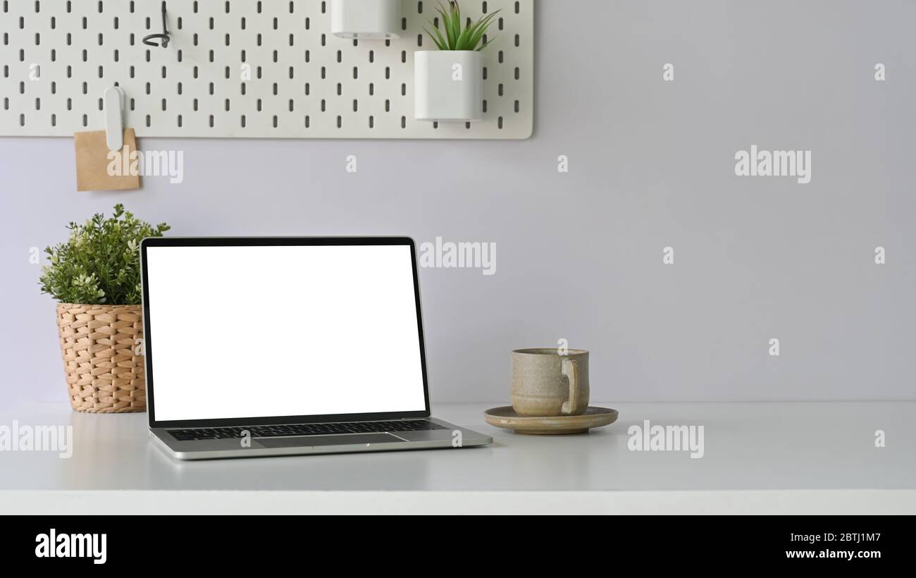 Foto di computer portatile bianco schermo vuoto mettere su scrivania bianca che circondato da caffè tazza e pianta in vimini cesto sopra comodo si Foto Stock