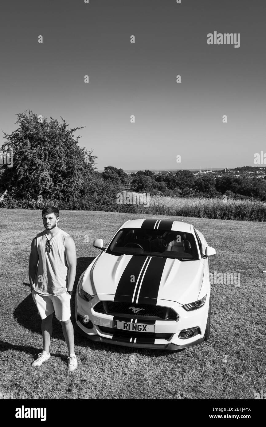 Un giovane muscoloso si pone con la sua auto sportiva Ford Mustang, presa in bianco e nero Foto Stock