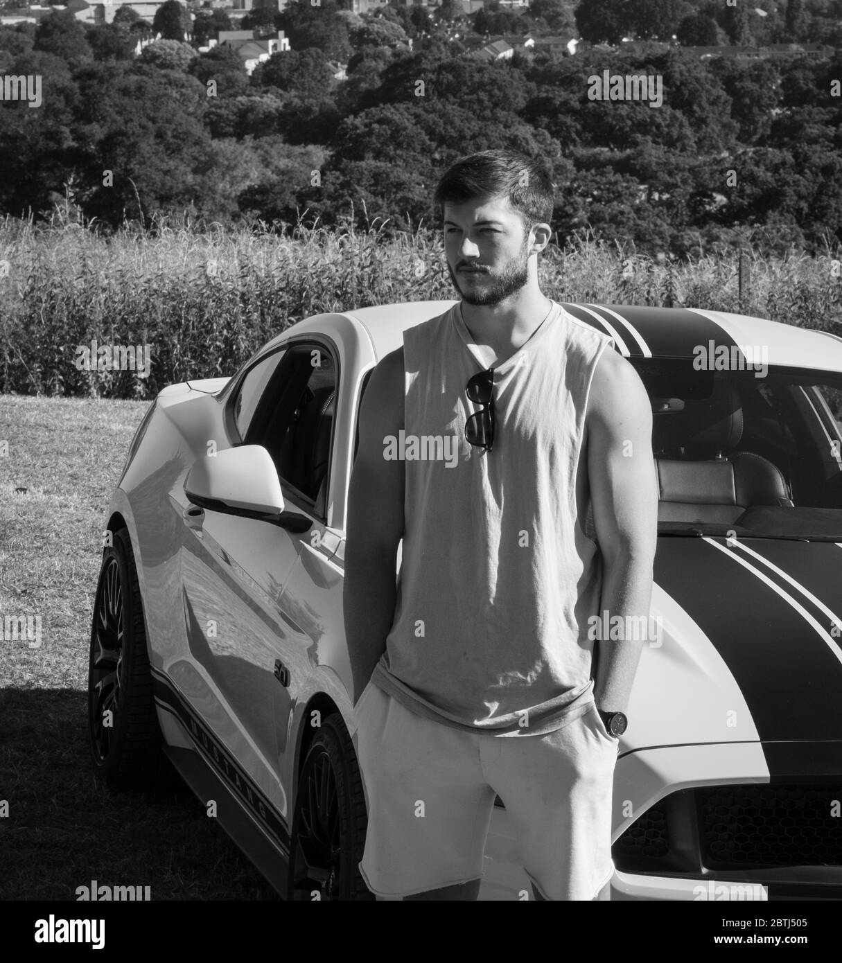 Un giovane muscoloso si pone con la sua auto sportiva Ford Mustang, presa in bianco e nero Foto Stock