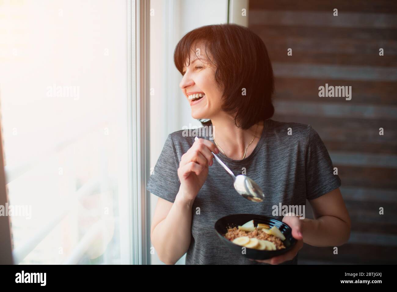 Donna adulta slim fit che mangia la colazione a casa. Prenditi cura della salute e del benessere. Persona anziana che ha la colazione in ciotola. Guardando Foto Stock
