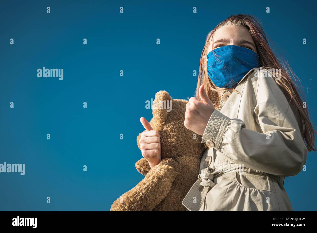 Tema Pandemia di Covid 19. Ragazza caucasica in maschera blu tenendo grande orsetto che mostra le miniature durante l'epidemia globale di virus. Foto Stock