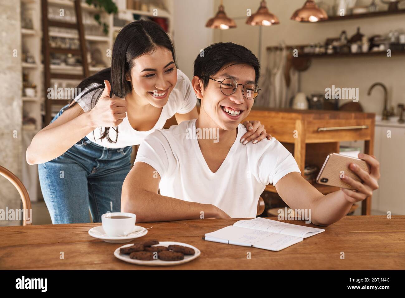 Immagine di una coppia sorridente multiculturale scattando foto selfie sullo smartphone e gesturando il pollice in su in cucina accogliente Foto Stock