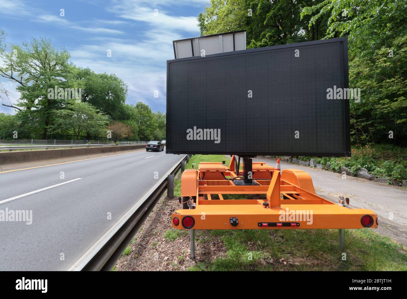 un cartello digitale vuoto per le spalle dell'autostrada, per lo spazio di copia, con la strada su un lato e una macchina che si allontana in lontananza, circondata da fogliame. Foto Stock