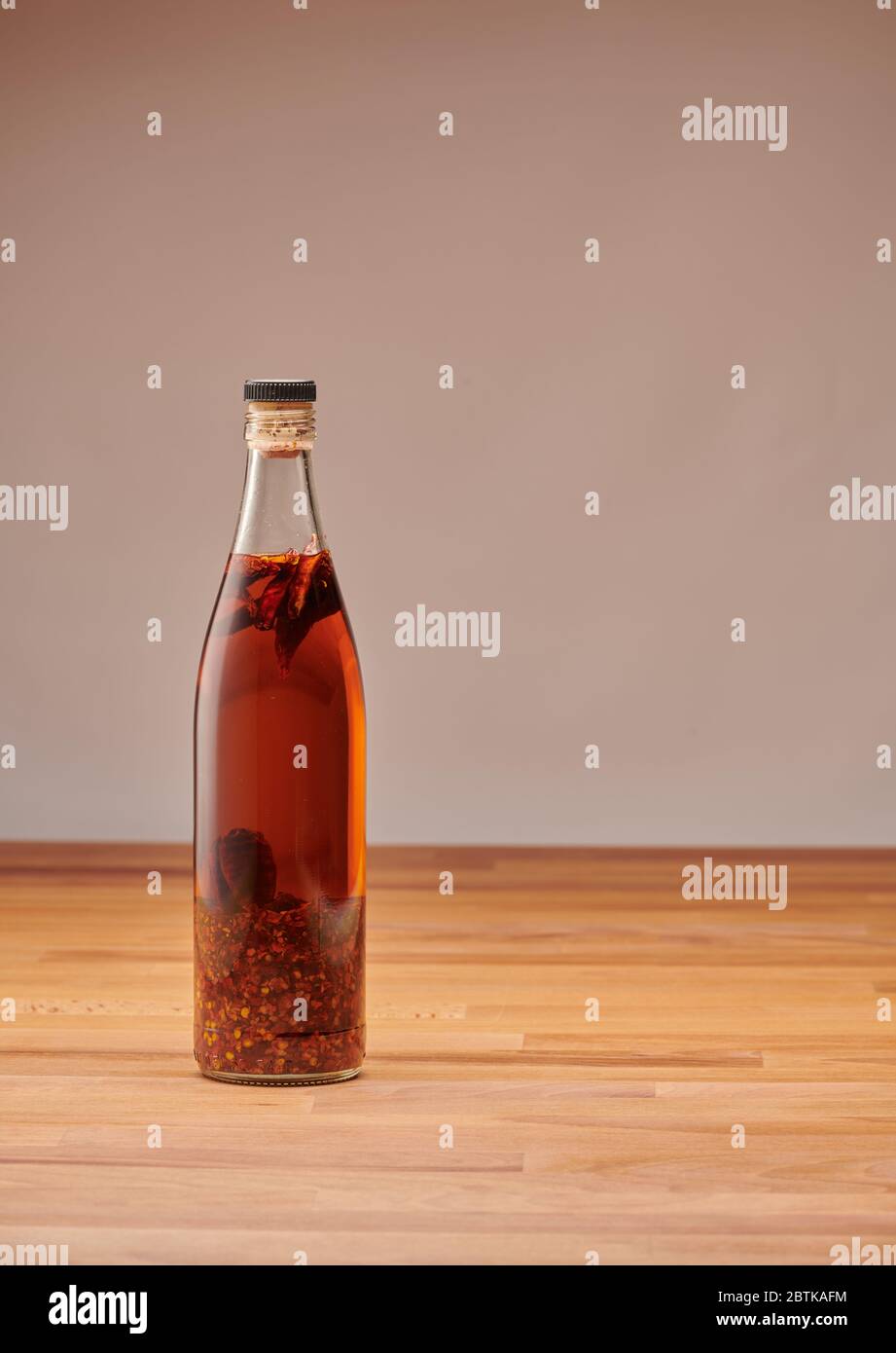 Bottiglia trasparente di olio di peperoncino fatto in casa con pezzi di peperoncino secco, su un tavolo di legno Foto Stock
