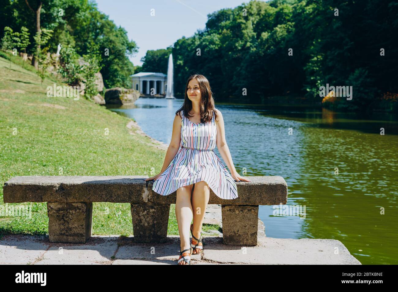 Sofia Park, Uman. Giovane donna seduta su una panchina sullo sfondo del lago con una fontana. La ragazza in un vestito si siede su una panchina di pietra. Foto Stock