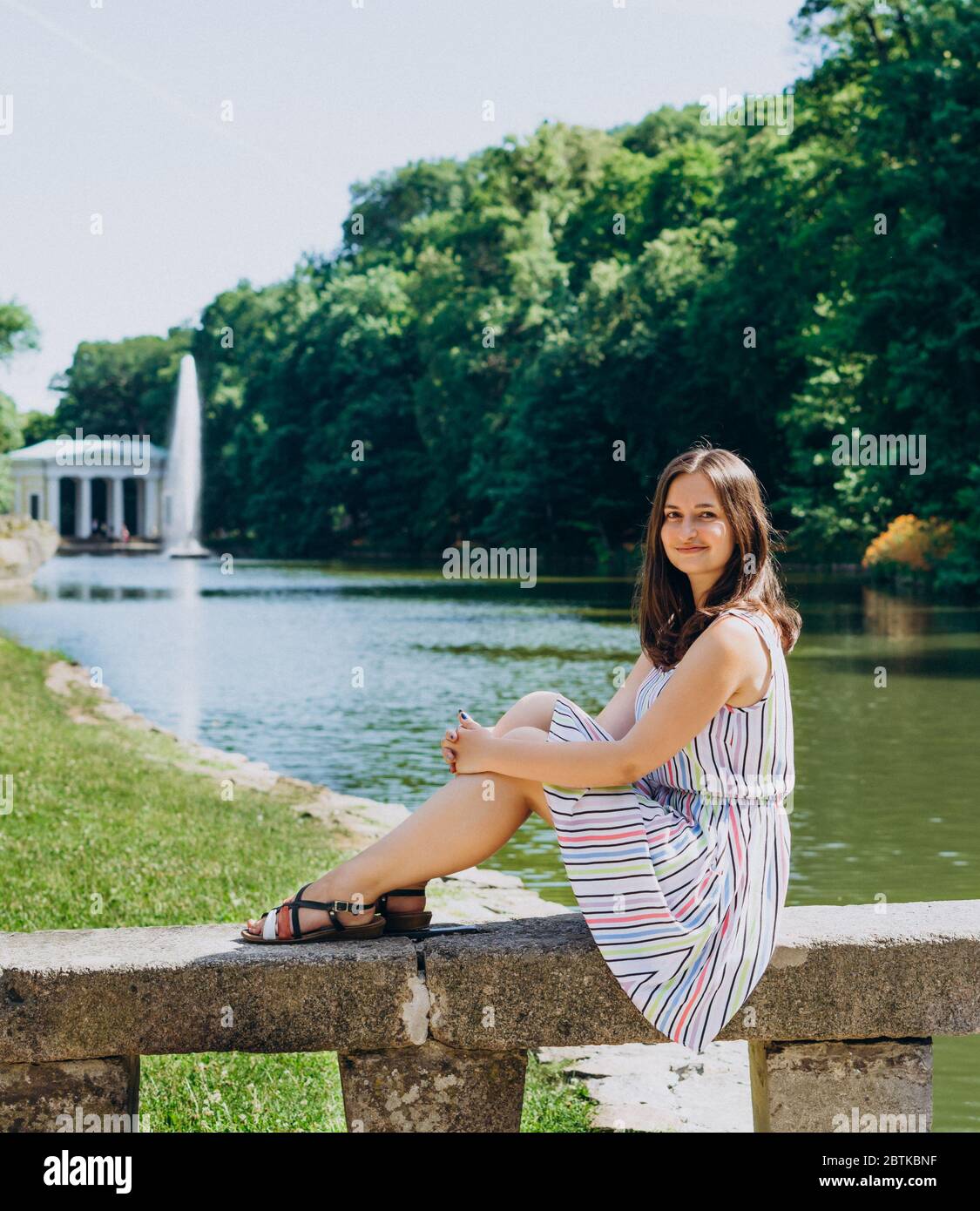 Sofia Park, Uman. Giovane donna seduta su una panchina sullo sfondo del lago con una fontana. La ragazza in un vestito si siede su una panchina di pietra. Foto Stock