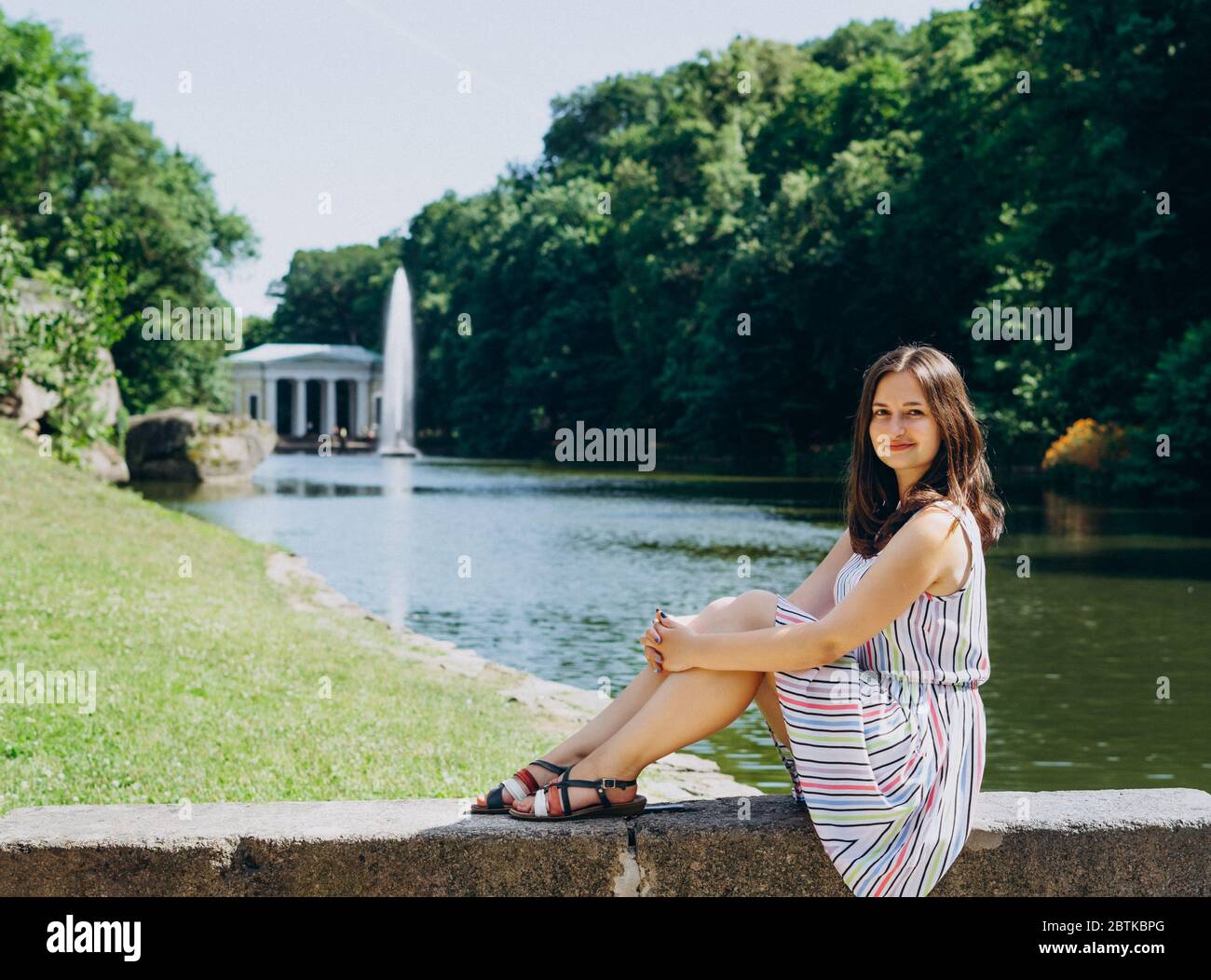 Sofia Park, Uman. Giovane donna seduta su una panchina sullo sfondo del lago con una fontana. La ragazza in un vestito si siede su una panchina di pietra. Foto Stock