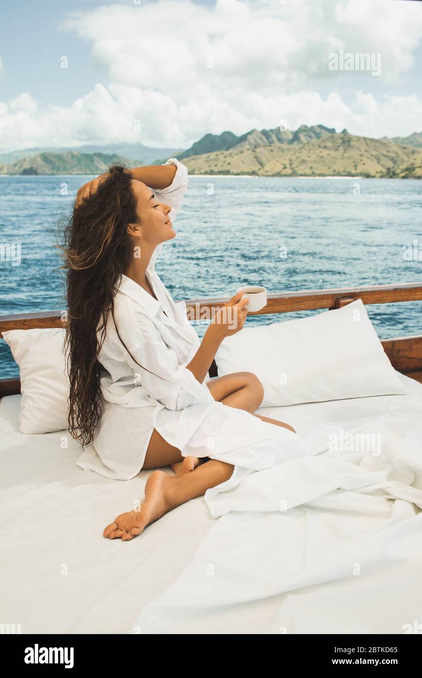 Donna che ama il caffè mattutino in barca in un tour privato in crociera. Letto a bordo. Viaggi e libertà di vita. Vista sulle montagne del parco nazionale di Komodo all'orizzonte. Foto Stock