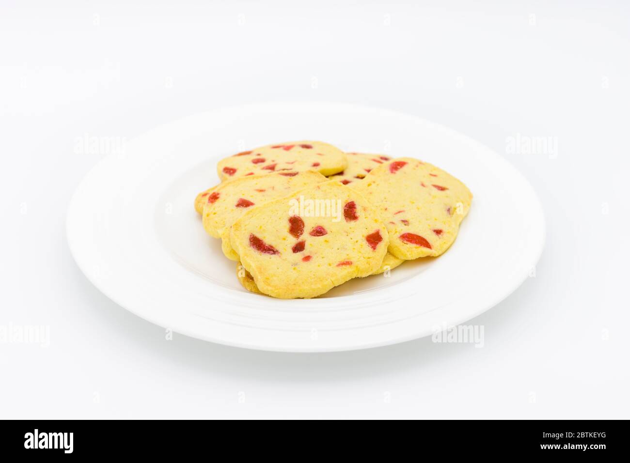 Un mucchio di biscotti tradizionali croccanti a fette e da cuocere, biscotti dorati fatti in casa con pezzi di ciliegia rossa presentati su un piatto bianco Foto Stock