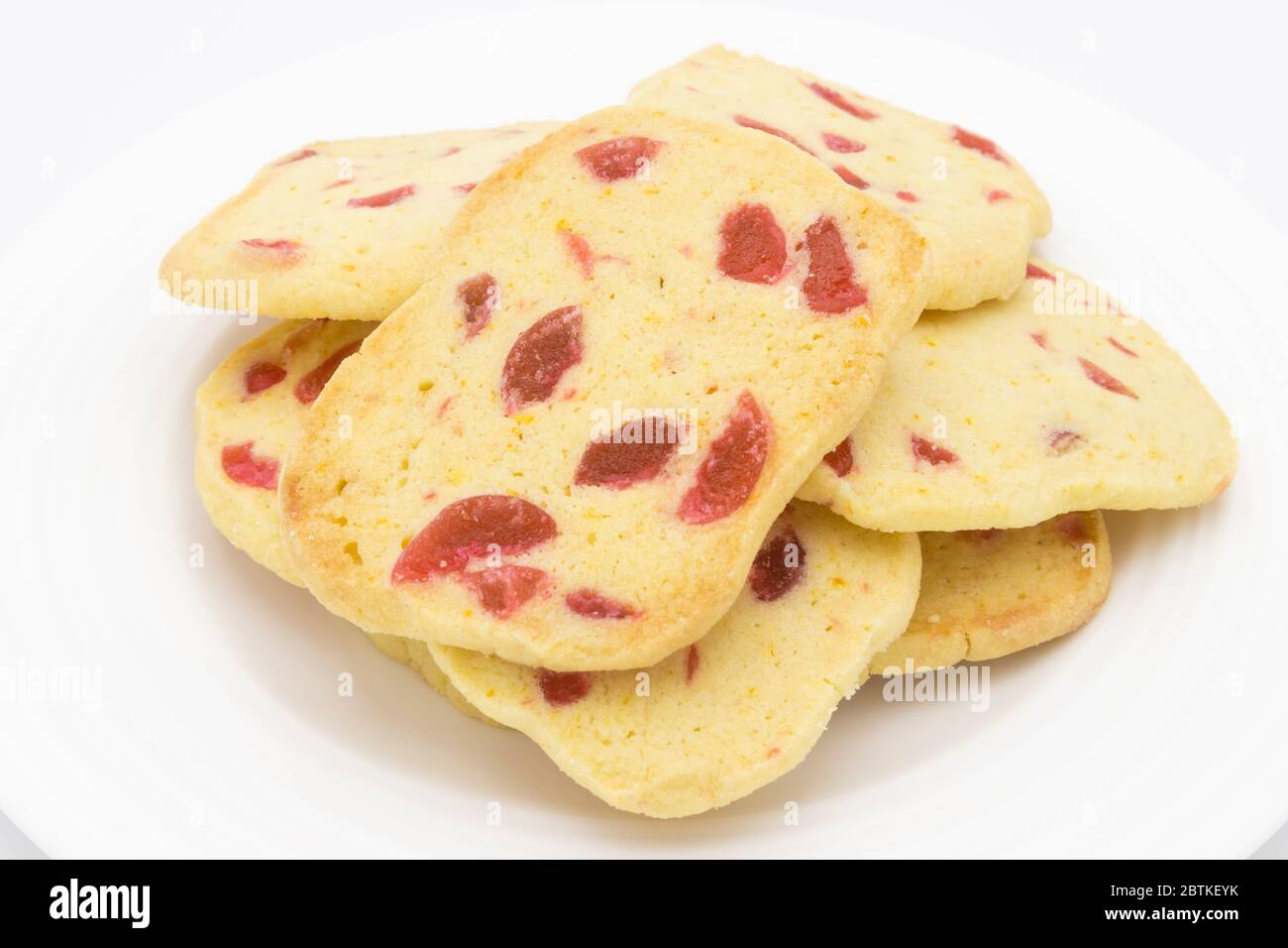 Un mucchio di biscotti tradizionali croccanti a fette e da cuocere, biscotti dorati fatti in casa con pezzi di ciliegia rossa presentati su un piatto bianco Foto Stock