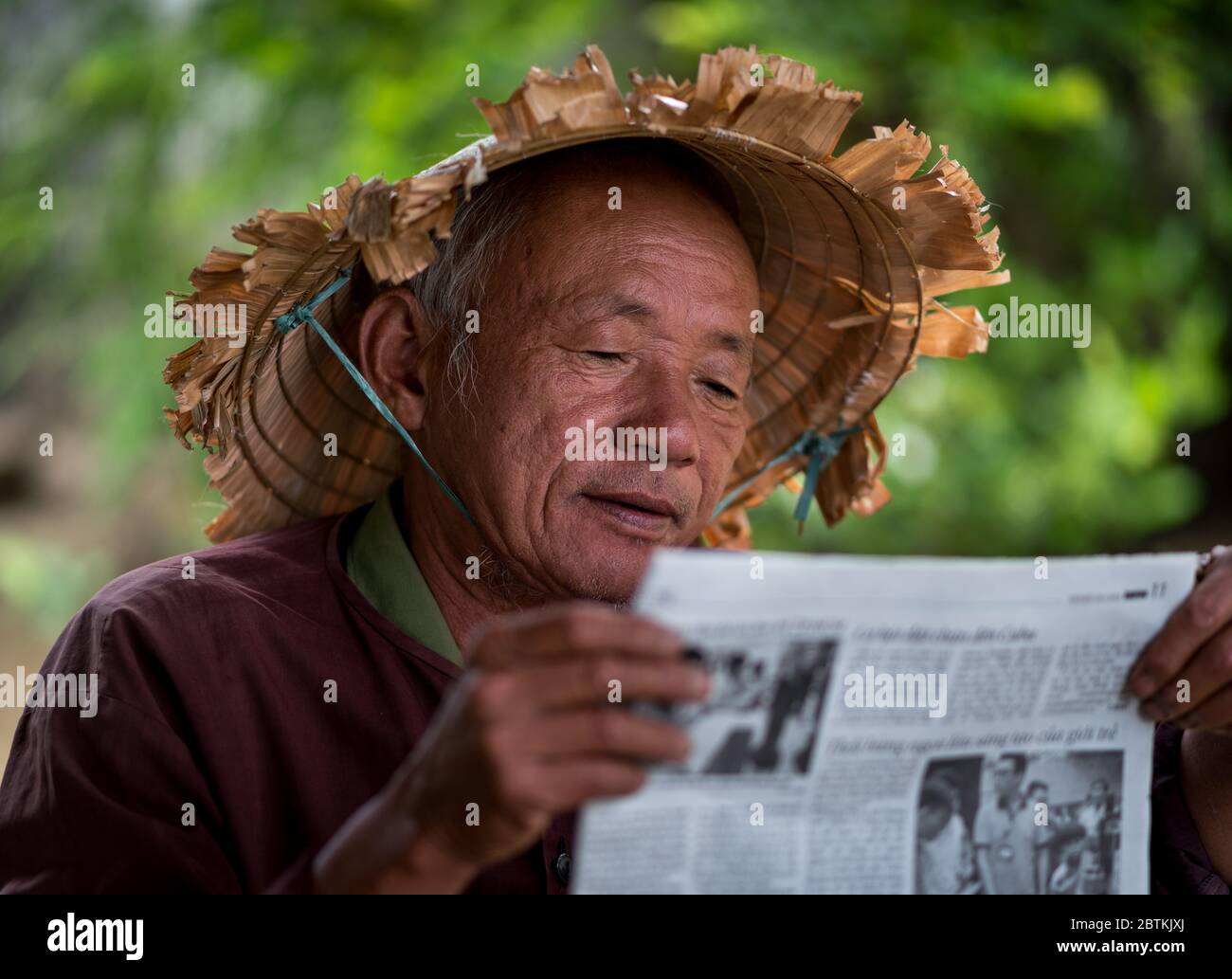 HOI AN - 10 MARZO 2018: Un vecchio con un cappello asiatico conico leggere giornale all'aperto sotto una strada in Hoi An, Vietnam il 10 marzo 2018 Foto Stock