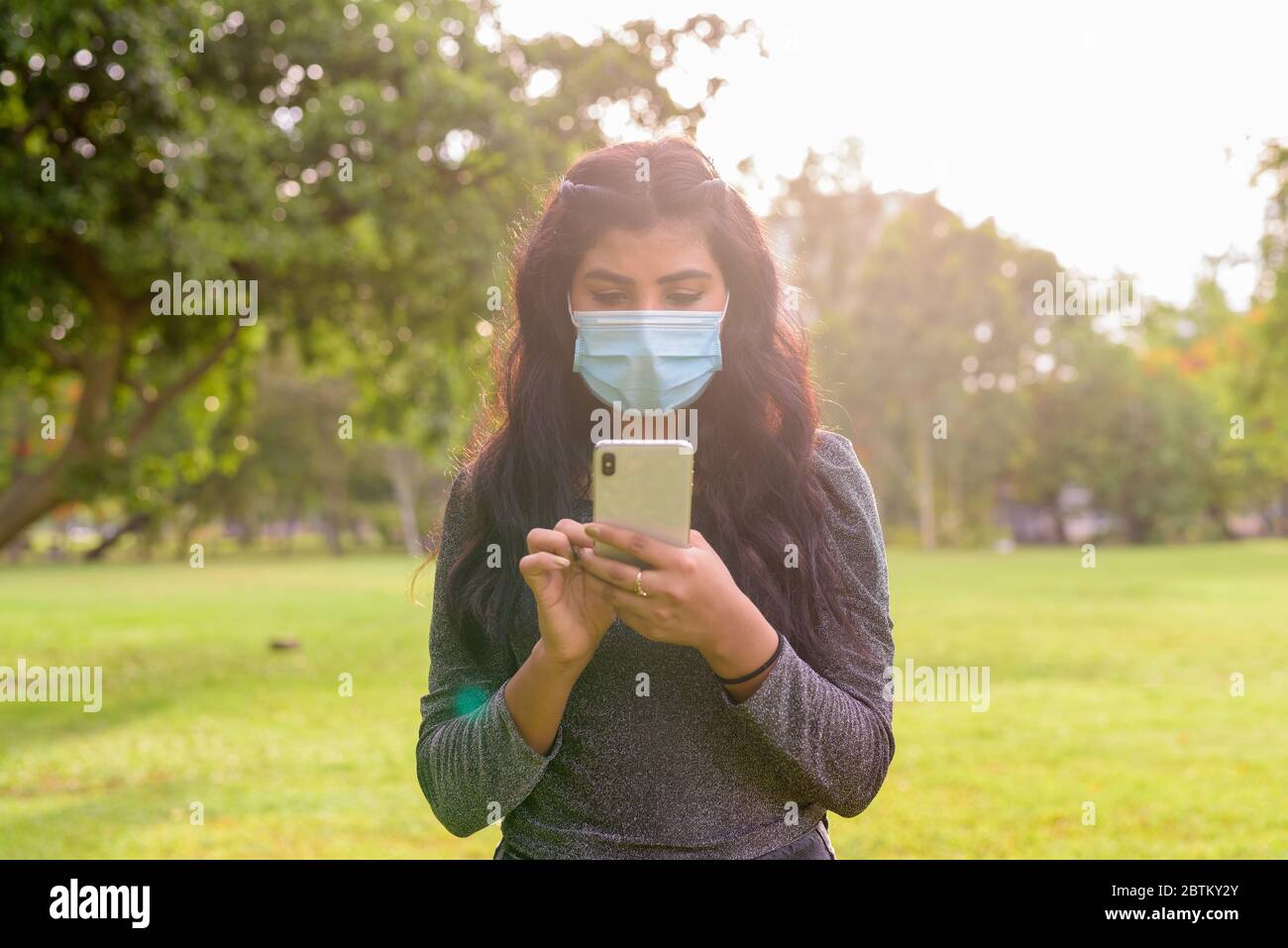 Giovane donna indiana con maschera utilizzando il telefono al parco Foto Stock