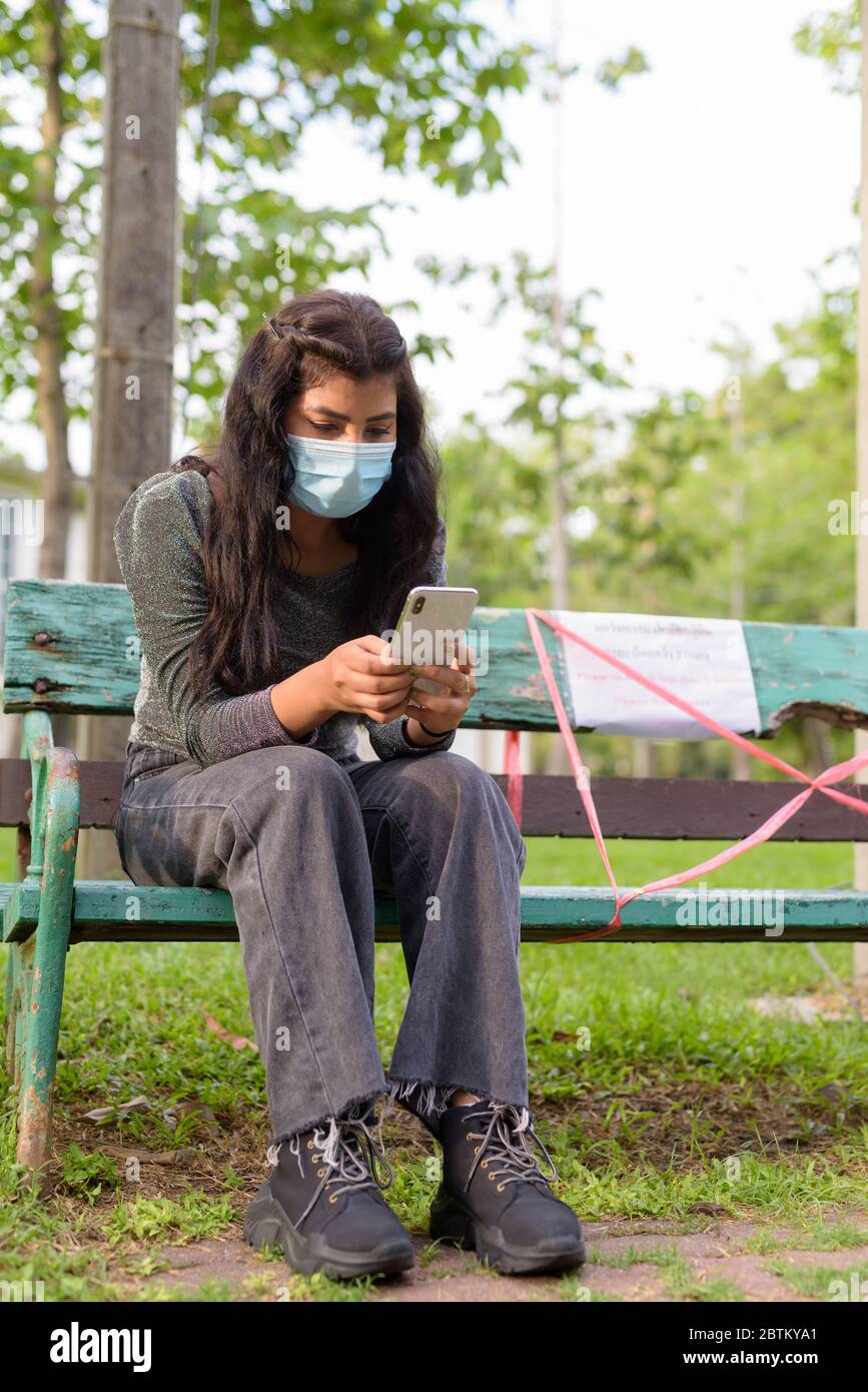 Giovane donna indiana con maschera usando il telefono mentre si siede a distanza sul banco del parco Foto Stock