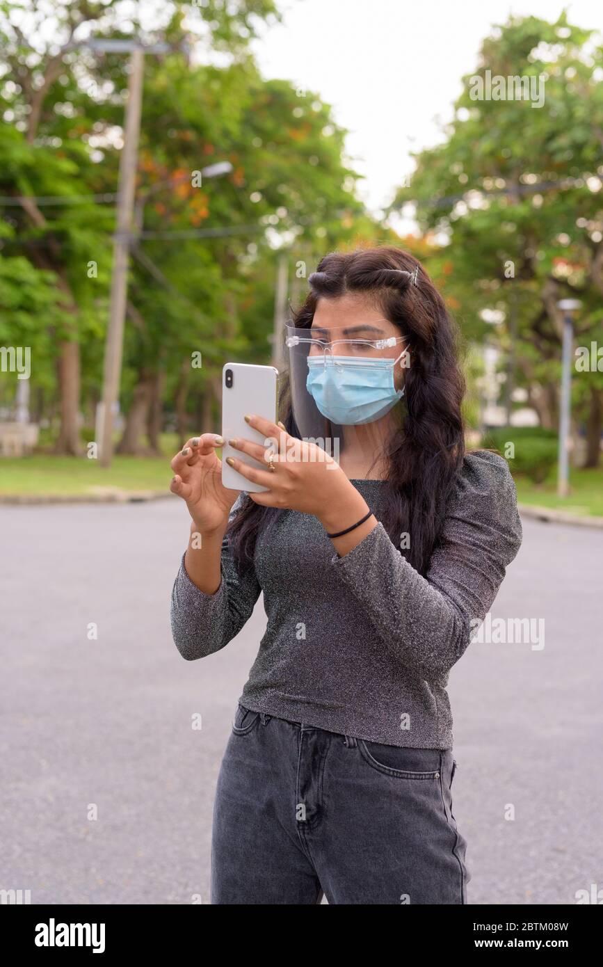 Giovane donna indiana con maschera e visiera che usa il telefono al parco all'aperto Foto Stock