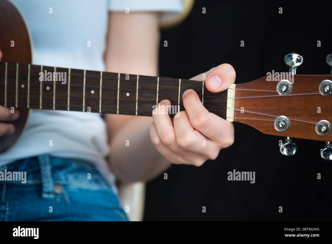 Primo piano della mano della ragazza che gioca G Major Chord sul tenore ukulele mentre seduto, con sfondo nero e spazio di copia Foto Stock