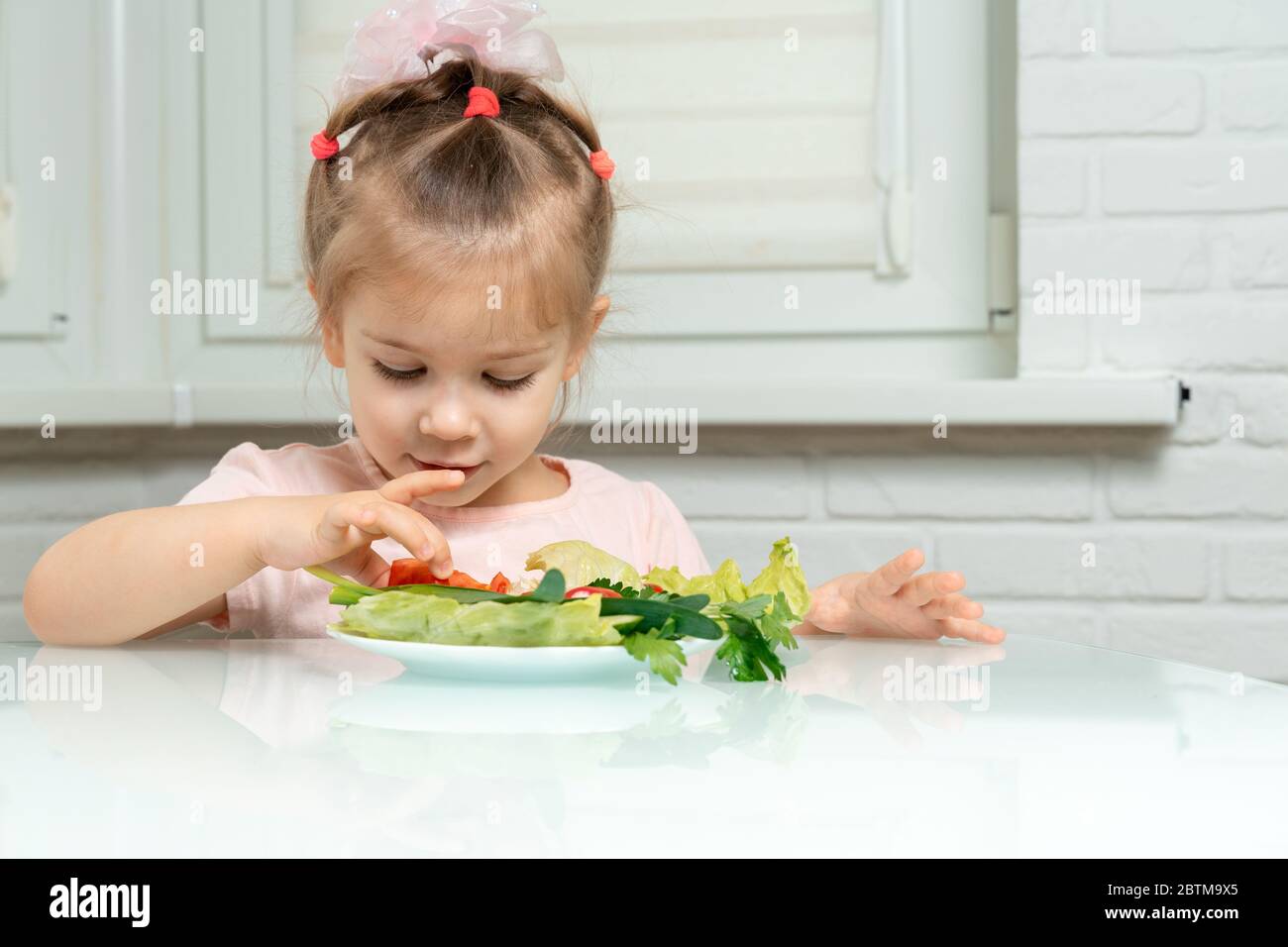 la ragazza di 4 anni si siede in cucina al tavolo e mangia le verdure, prende un pomodoro tritato da un piatto. regole nutrizionali prescolare Foto Stock
