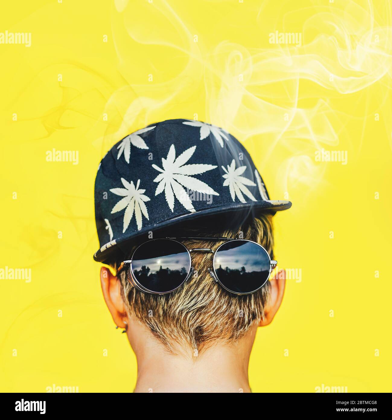 Giovane ragazza in occhiali da sole e cappellino da baseball con foglie di marijuana fuma su uno sfondo giallo parete. Soffici di fumo di canapa Foto Stock