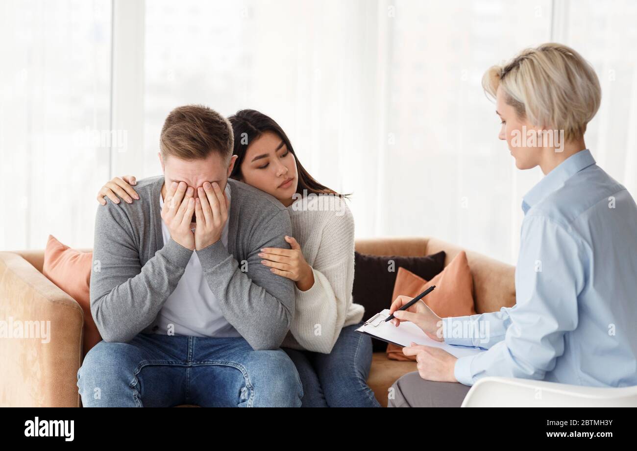 Moglie che conforta il marito piangente durante l'appuntamento di Psicoterapia nell'ufficio dello psicologo Foto Stock