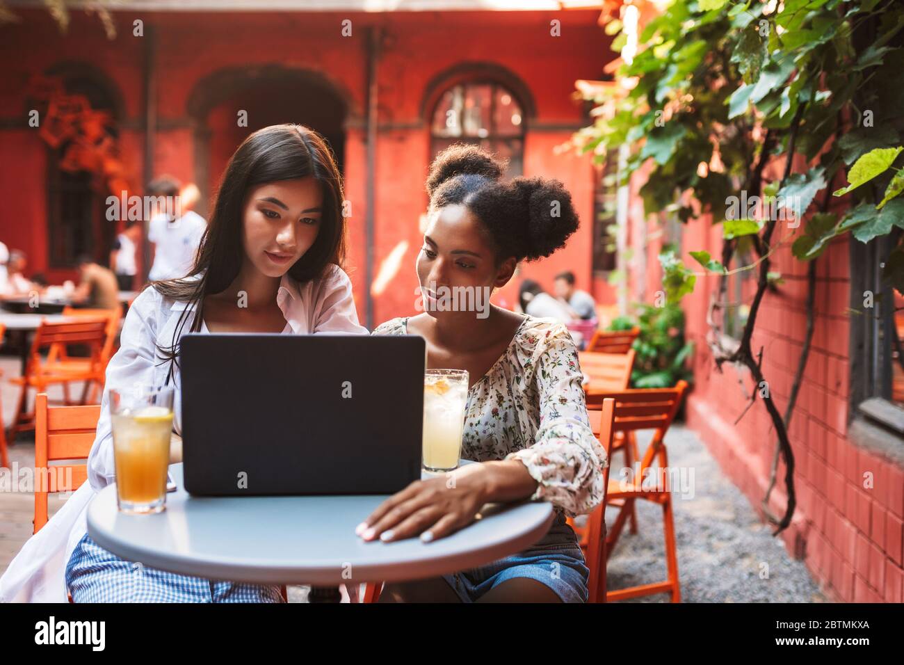 Due belle ragazze usano il computer portatile mentre si trascorrono dei cocktail insieme nell'accogliente cortile della caffetteria Foto Stock