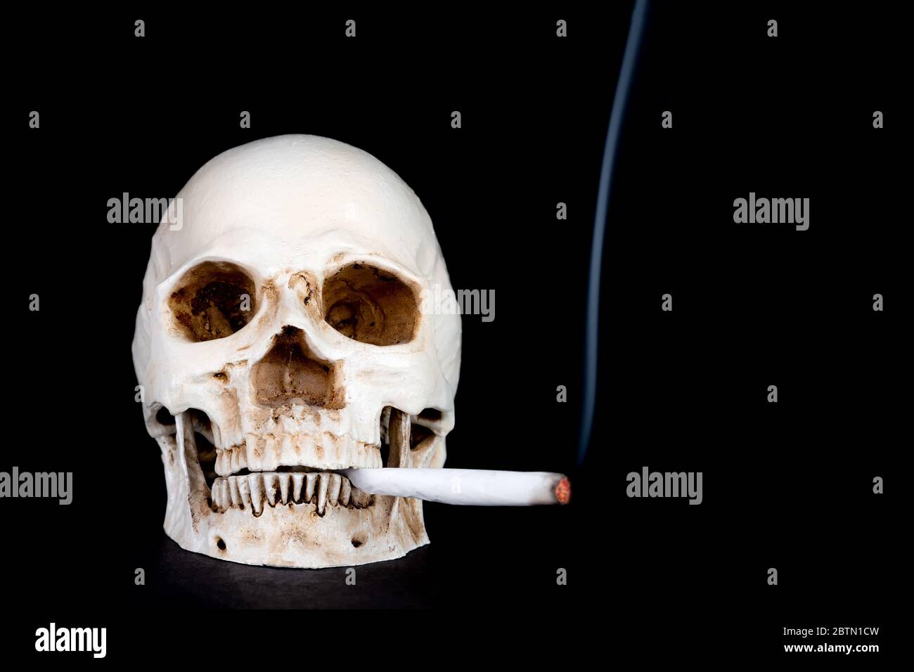 Un cranio umano che fuma e guarda direttamente allo spettatore, fumando una sigaretta di cannabis arrotolata a mano. Sottile linea di fumo. Isolato su sfondo nero. Foto Stock