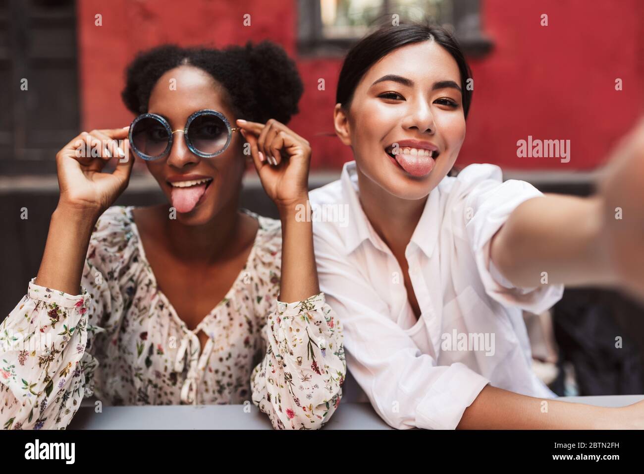 Due graziose ragazze che mostrano felicemente le lingue mentre scattano le foto insieme che trascorrono il tempo nel cortile del caffè Foto Stock