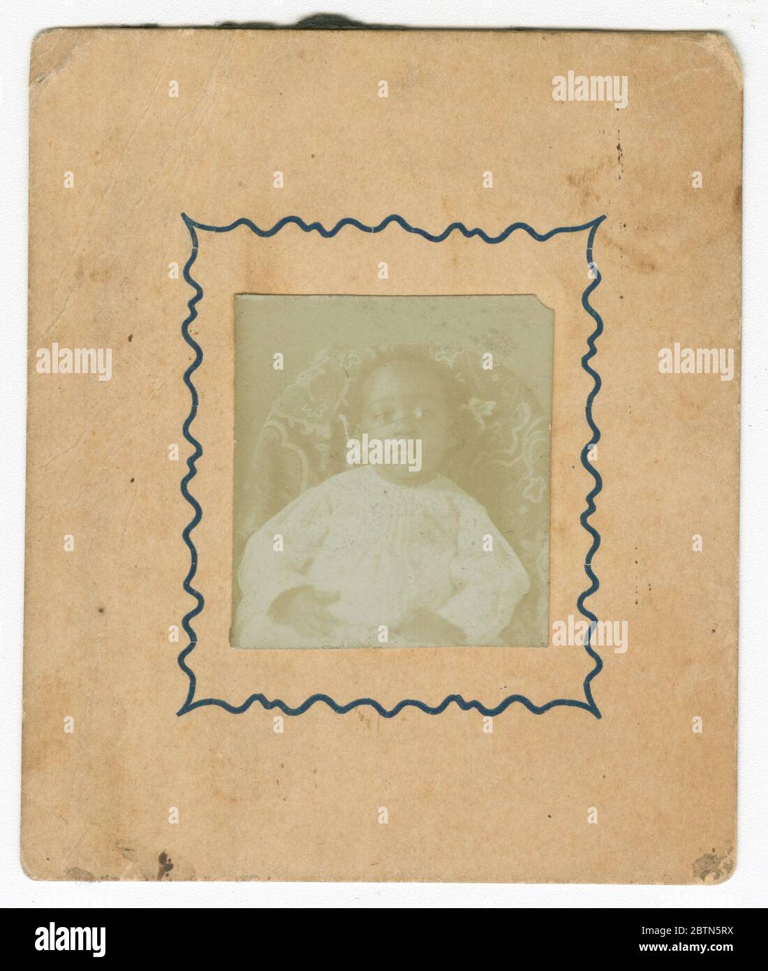 Fotografia di un bambino vestito di bianco e seduto su una sedia. Un ritratto di un bambino vestito di bianco con cartes-de-visite dai toni seppia. Il bambino è seduto su una sedia di colore scuro con un design decorativo di colore chiaro su di essa. La fotografia è delimitata da una linea nera ondulata. Non sono presenti iscrizioni sulla parte anteriore o posteriore. Foto Stock