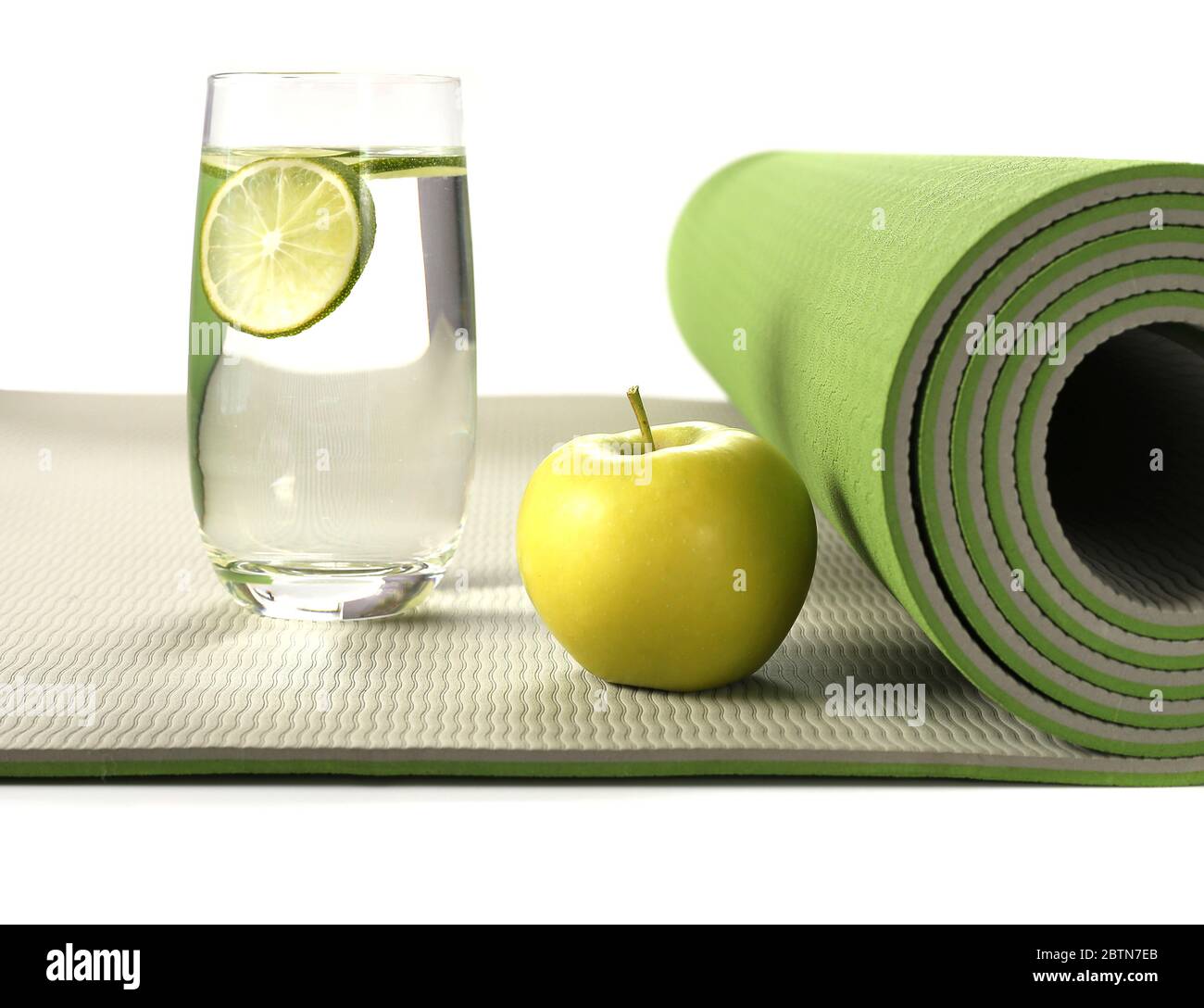 Tappeto yoga verde con mela e bicchiere d'acqua con limone su sfondo bianco, concetto di vita sana Foto Stock