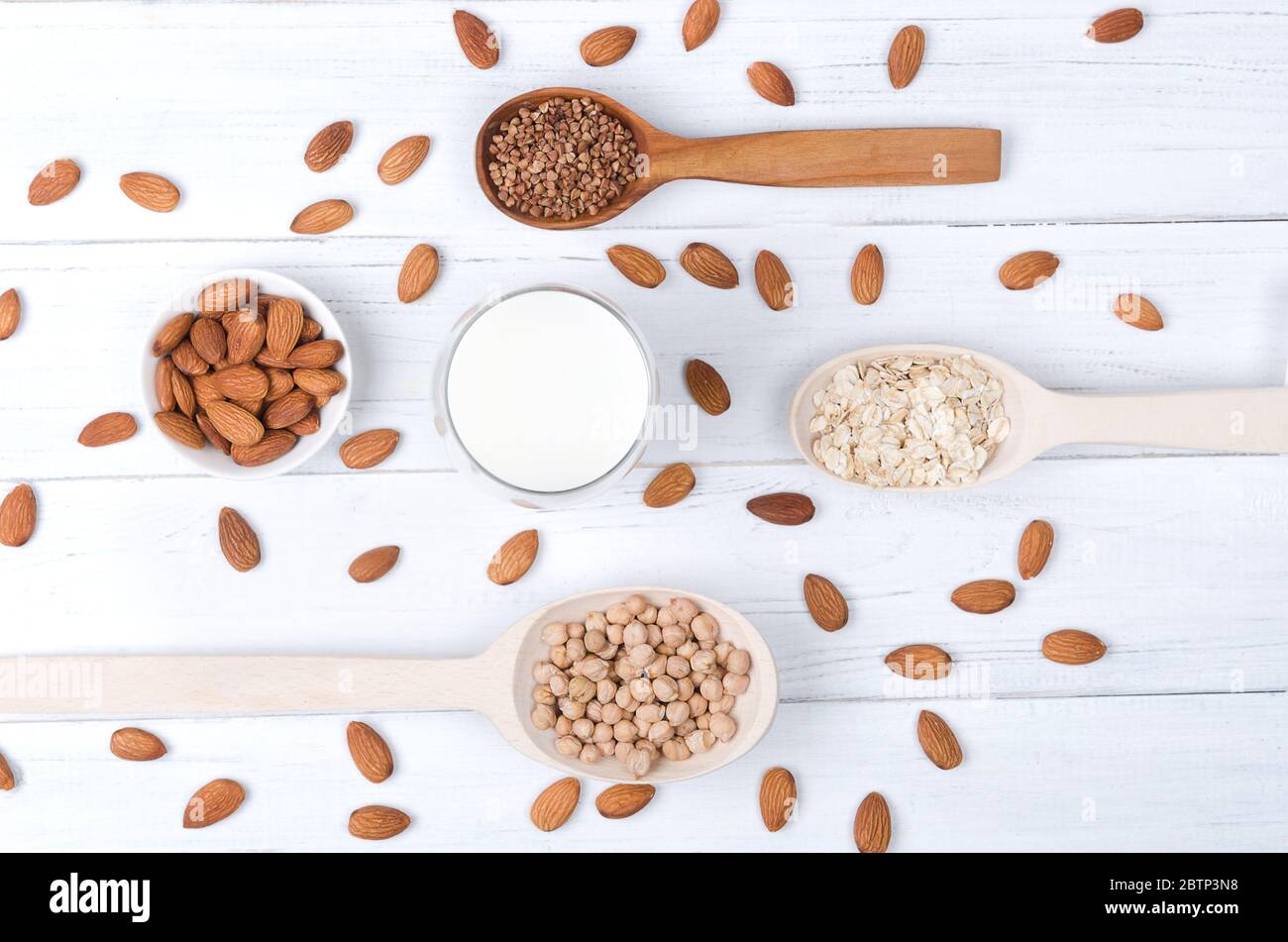 Vista su piatto del latte di mandorle in vetro con semi di mandorle, fibbietta, avena e cucchiai di legno su tavolo di legno bianco Foto Stock