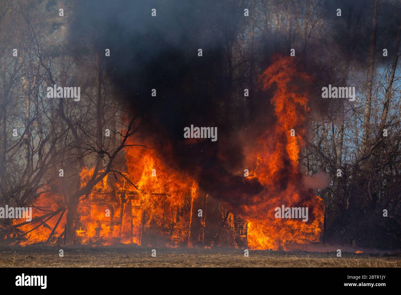 Fumo nero e fiamme che sparano da un vecchio edificio circondato da alberi in un paesaggio rurale estivo Foto Stock