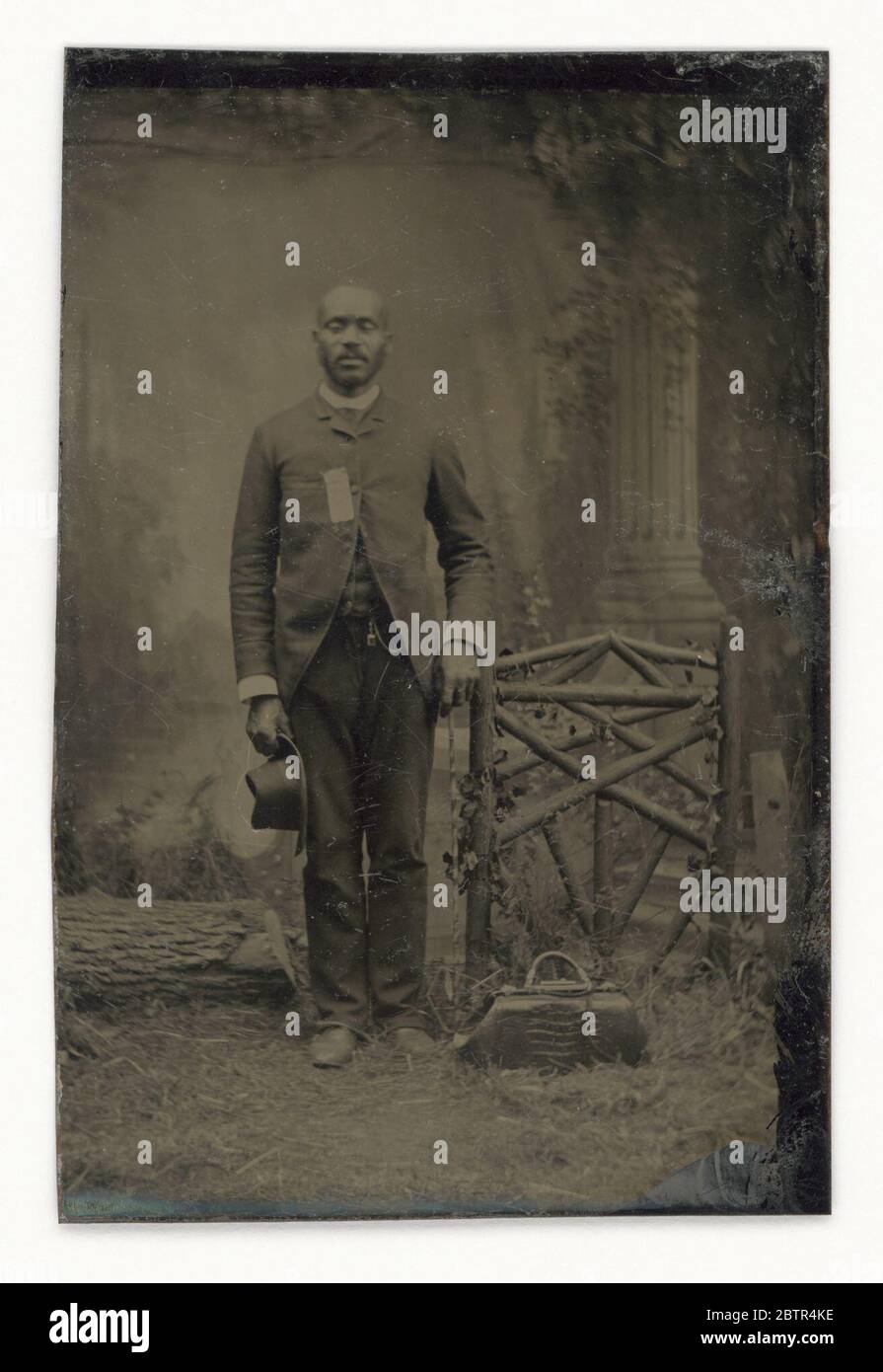 Tintype di un uomo. Una tintype di un uomo afro-americano non identificato in uno studio circondato da oggetti all'aperto e da un ambiente classico sullo sfondo. Tiene un cappello nella mano destra e una borsa si appoggia ai piedi. Foto Stock Tintype di un uomo. Una tintype di un uomo afro-americano non identificato in uno studio circondato da oggetti all'aperto e da un ambiente classico sullo sfondo. Tiene un cappello nella mano destra e una borsa si appoggia ai piedi. Foto Stock