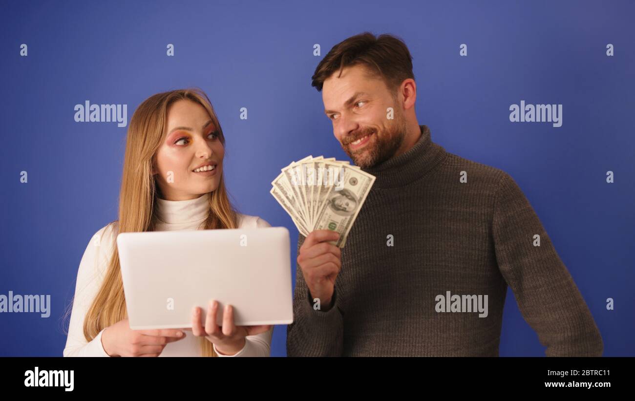 Giovane bella donna che mostra il conto sul tablet al suo ricco ragazzo che tiene in mano i contanti. Shopping online e rapporto con interesse. Foto Stock