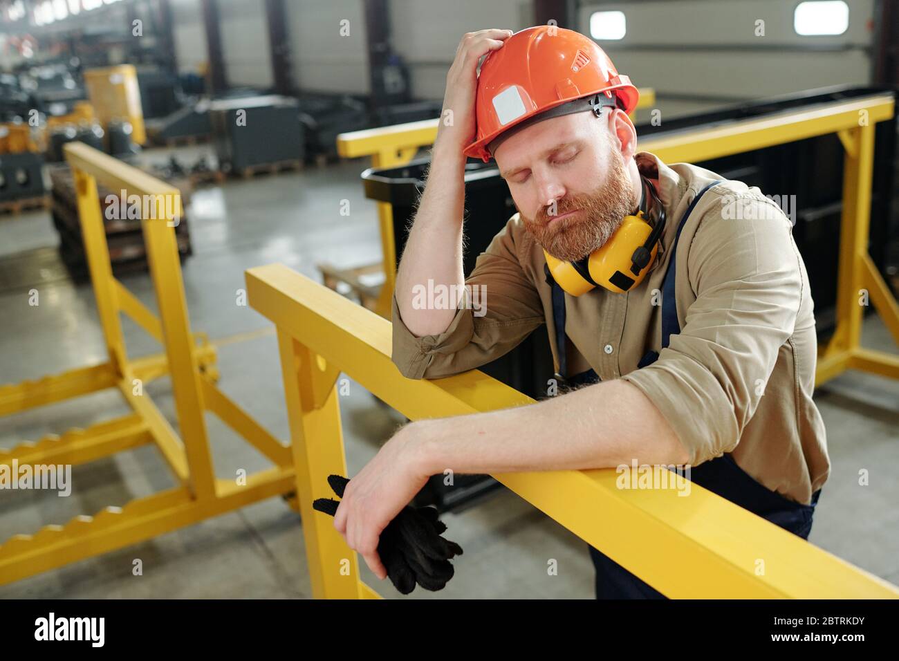 Perplesso operaio di fabbrica bearded che tiene gli occhi chiusi e appoggiandosi sulla ringhiera metallica gialla mentre pensa ai problemi di produzione alla fabbrica Foto Stock