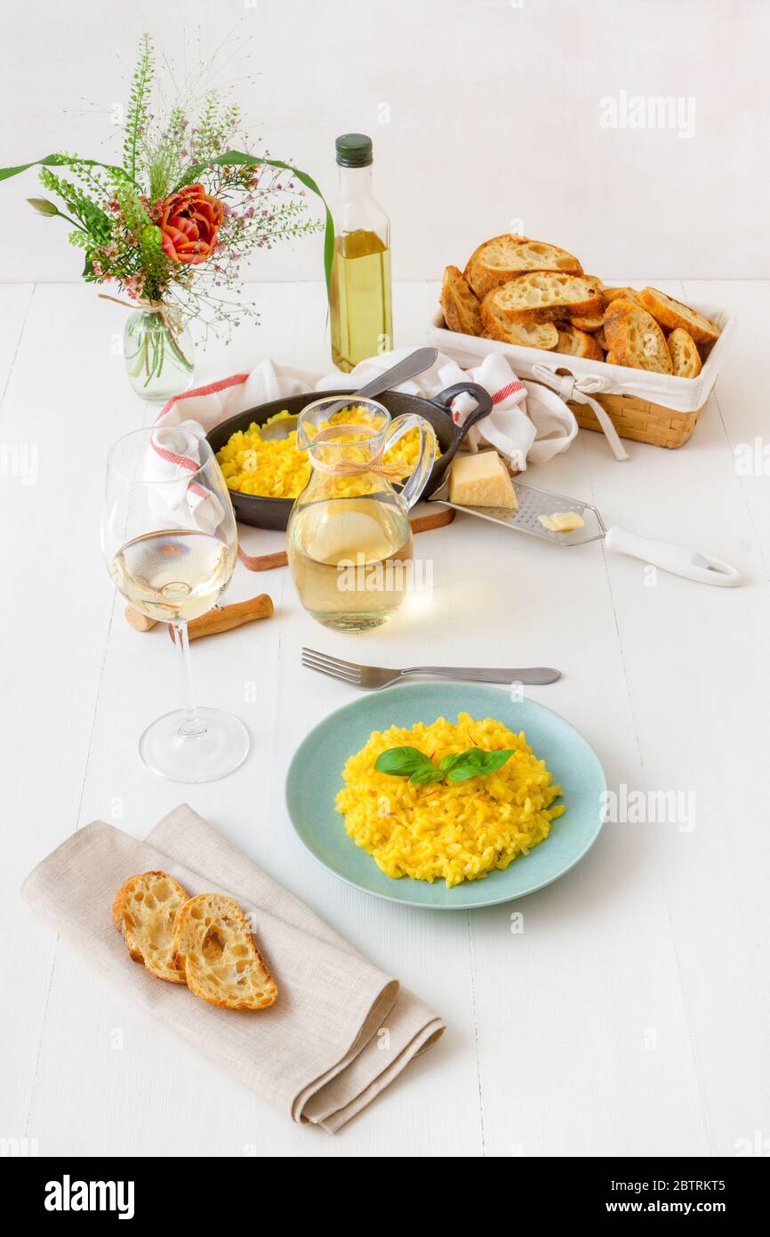 Risotto milanese, tavolo in legno con risotto tradizionale italiano allo zafferano, bicchieri e brocca di vino, bottiglia di olio d'oliva, cesto di pane e fiori Foto Stock