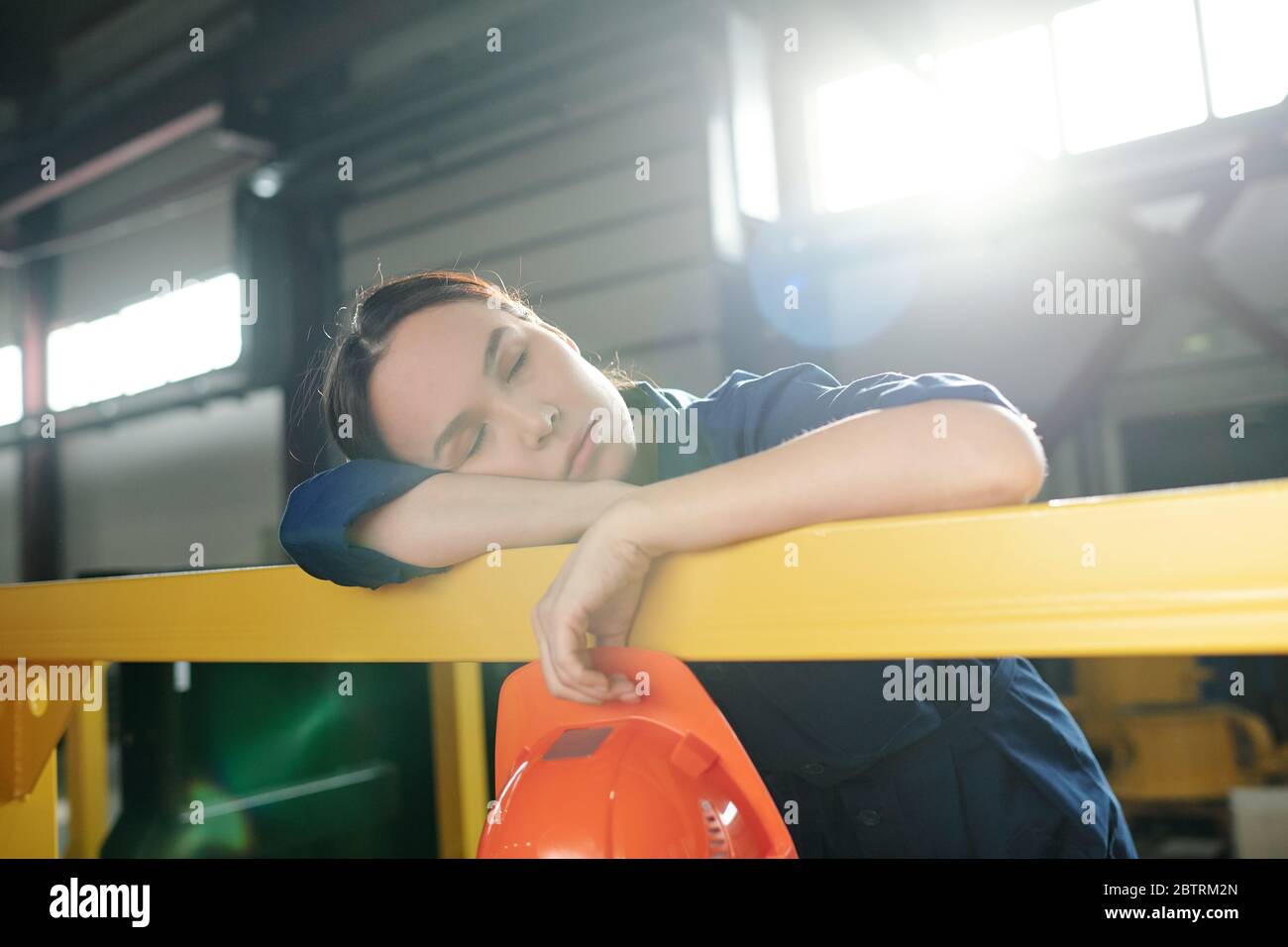 Stanco lavoratore asiatico dormire con l'hardHat in mano e appoggiarsi sulla ringhiera in fabbrica Foto Stock