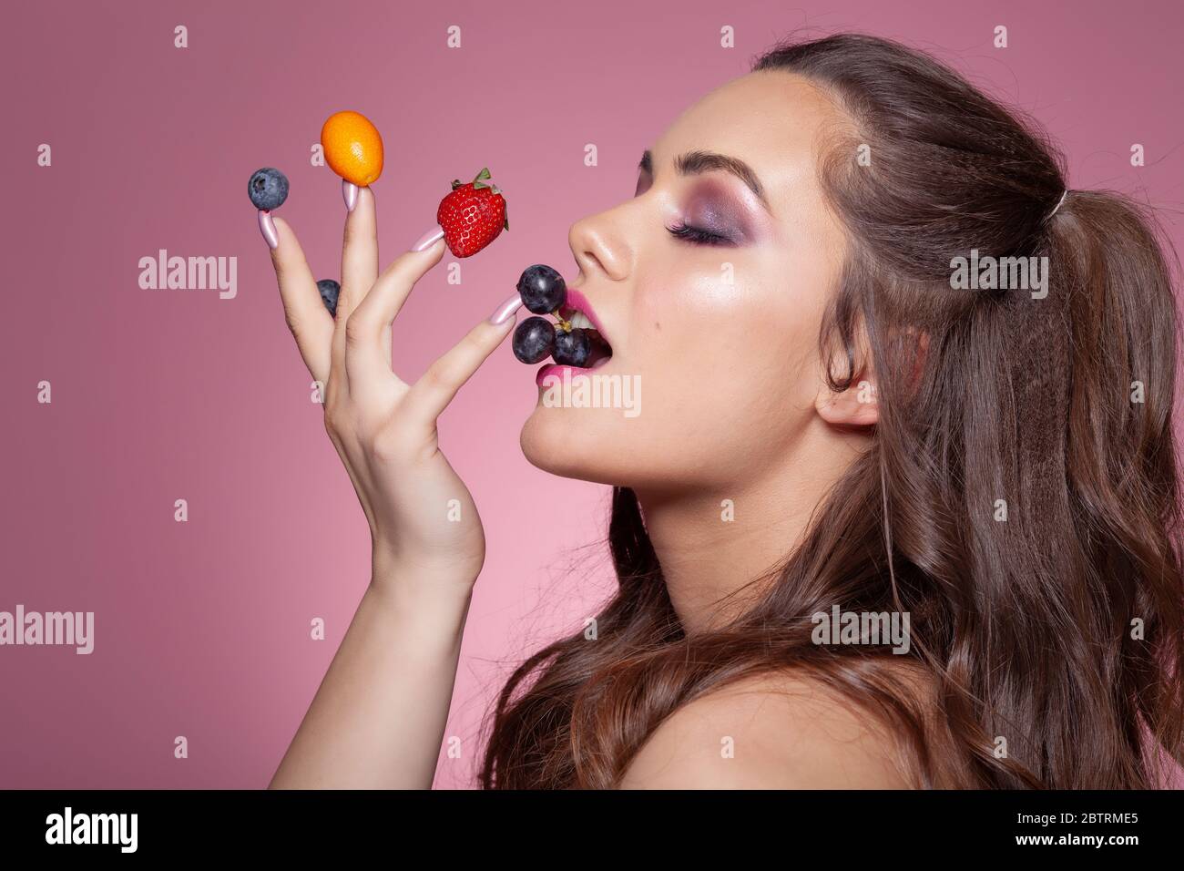 Giovane donna sexy che mangia frutta dalle dita. Riprese in studio su sfondo rosa. Foto Stock