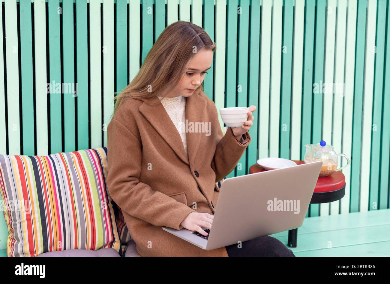 Attraente donna caucasica seduta da sola in un caffè all'aperto con uno sfondo verde luminoso. Giovane bella ragazza in un caffè che lavora su un computer portatile mentre si pranza e sorridendo. Foto Stock