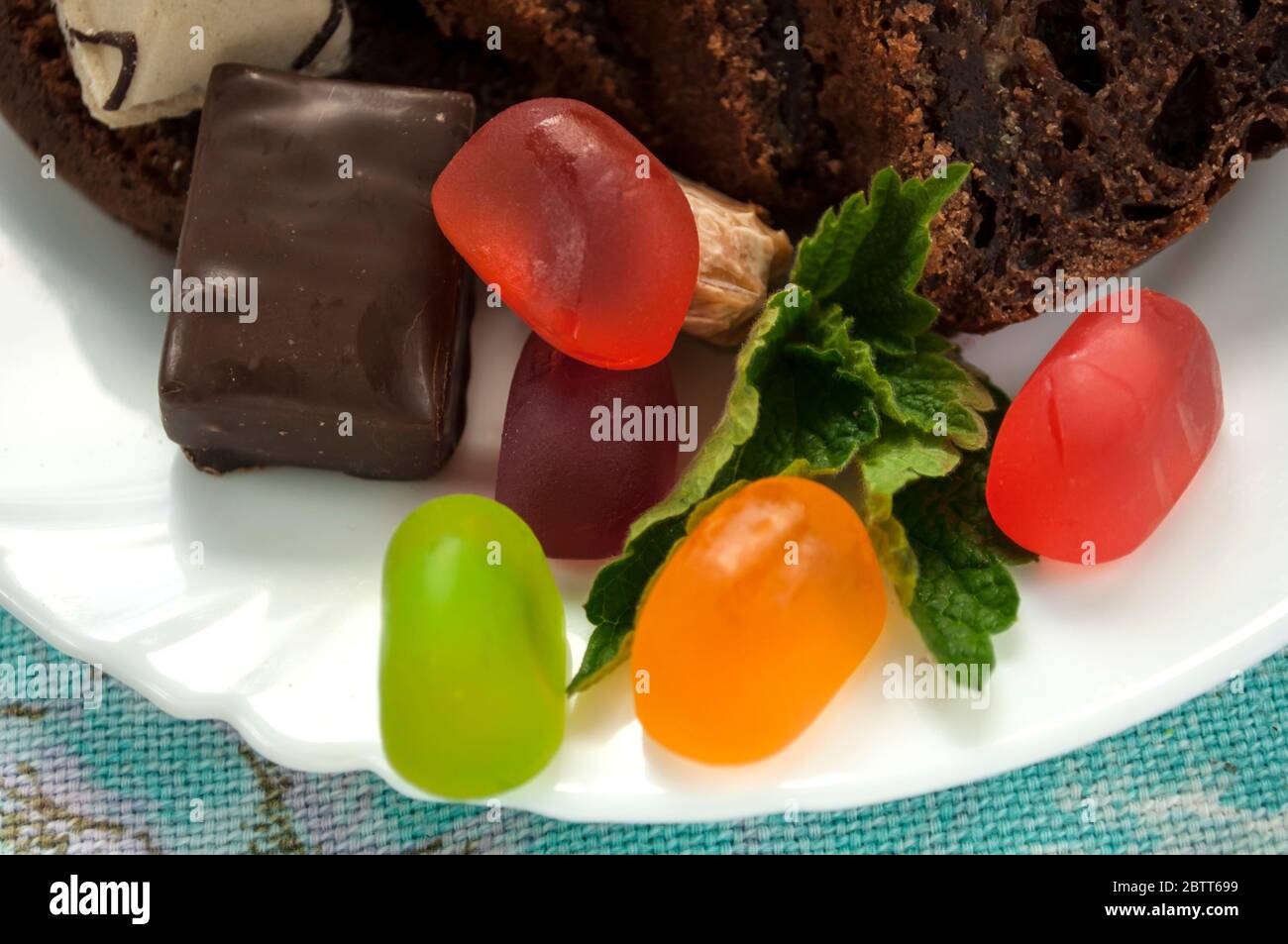 Pezzi di torta al cioccolato e caramelle di gelatina colorate su un piatto bianco decorato con primo piano di menta Foto Stock