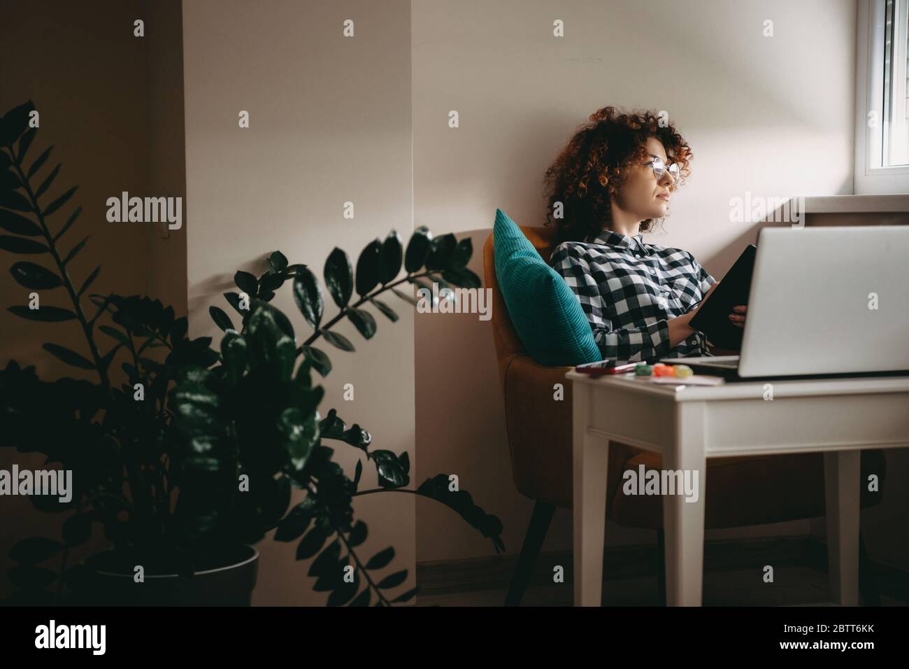 Giovane uomo d'affari con capelli ricci e occhiali che lavorano da casa al computer pensando a qualcosa mentre teneva un libro Foto Stock