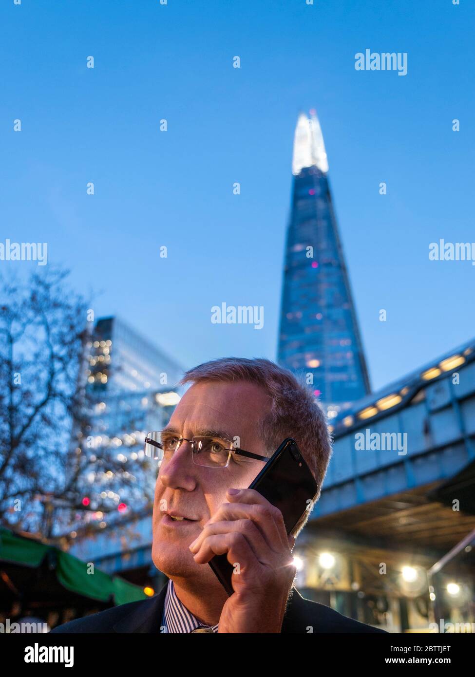 L'uomo d'affari di Shard City all'aperto parla sul suo smartphone nero al tramonto con la torre "The London Shard" e il treno metropolitano di Londra sfocato sullo sfondo Southwark London SE1 Foto Stock